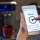 Hands holding smartphone connected to a smart coffeemaker, visible in background